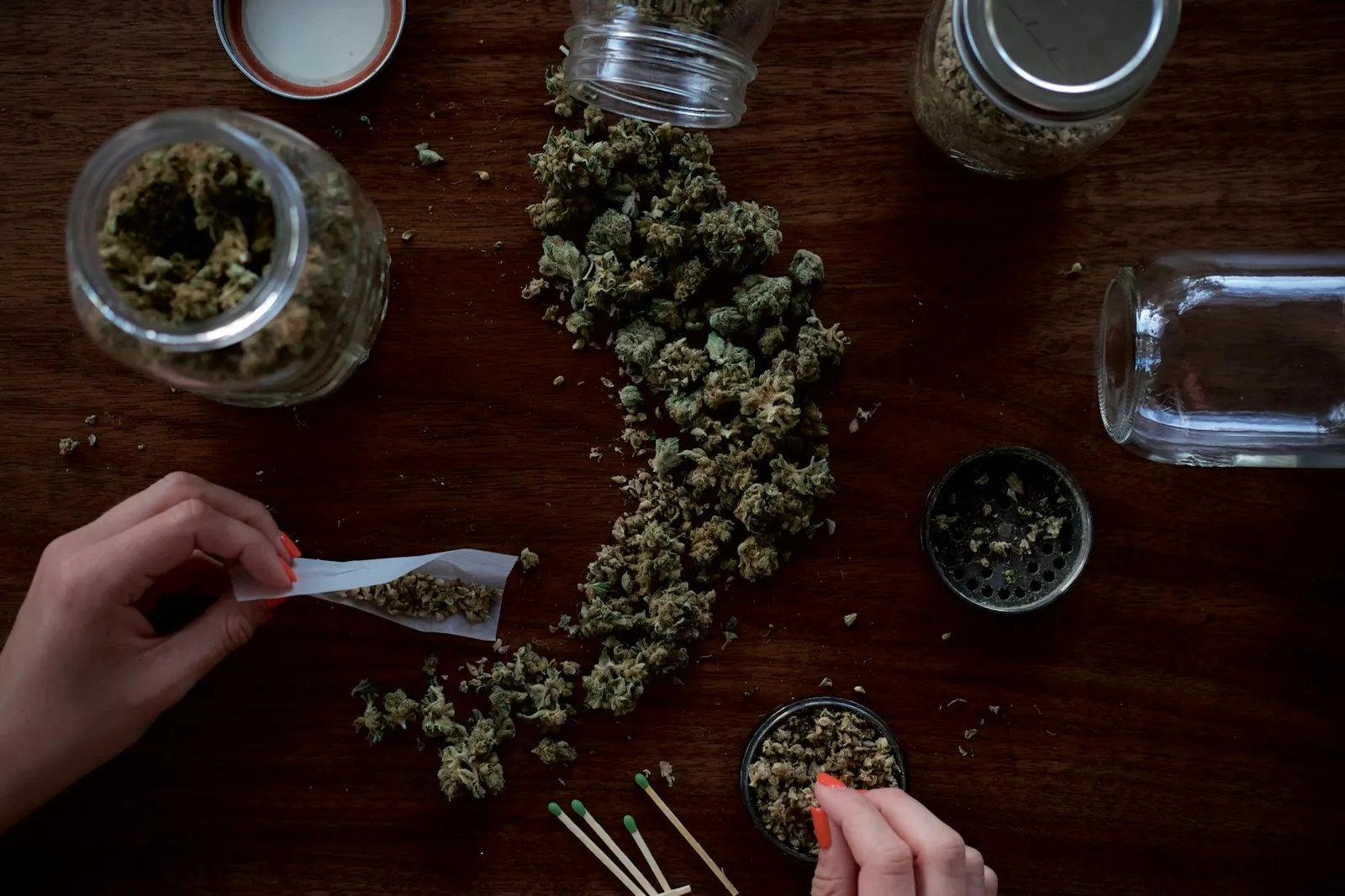 Cannabis buds, storage jars, and a joint being rolled on a wooden table